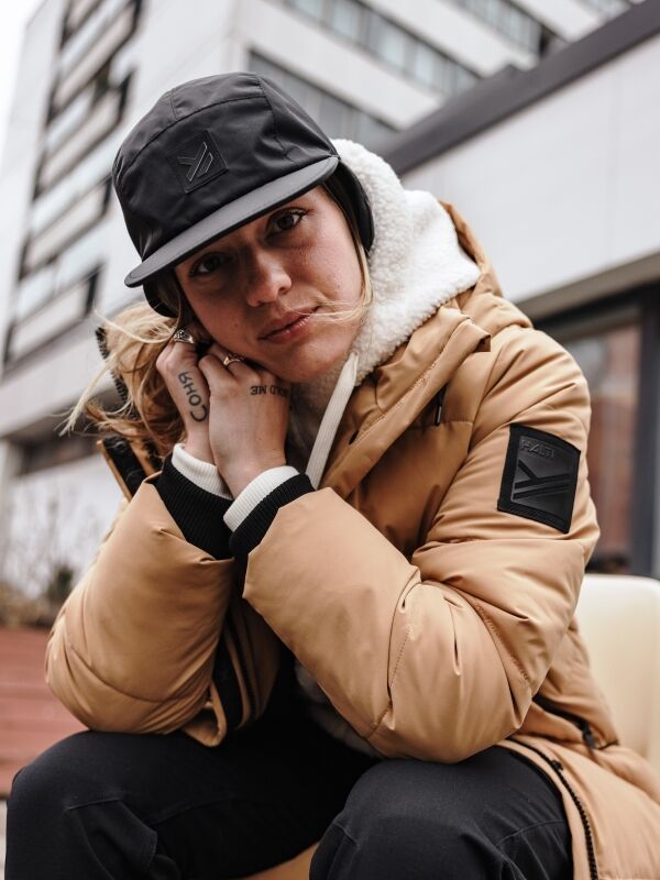 Eine Frau in einer hellbraunen „Halti Brahe“ Puffer-Langwinterjacke und einer schwarzen Mütze sitzt im Freien, im Hintergrund sind Stadtgebäude zu sehen, und sie blickt in die Kamera.