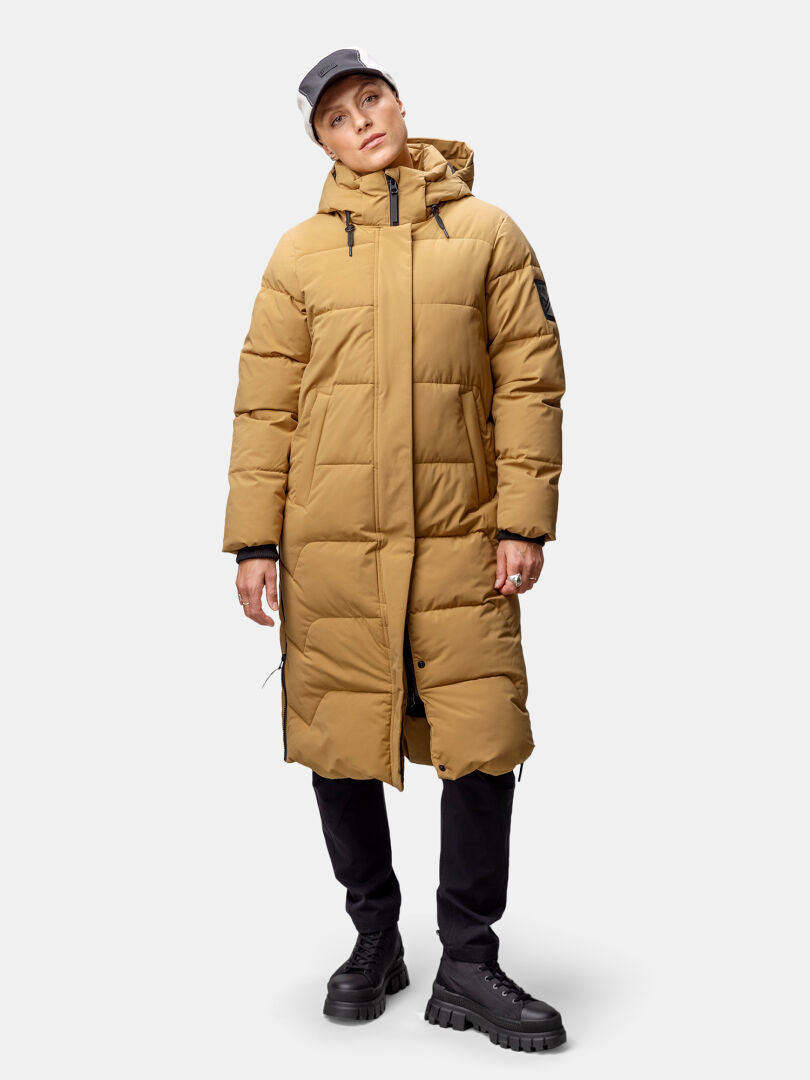 Eine Frau in einer langen Winterjacke von Halti Brahe, einer schwarzen Hose, klobigen Stiefeln und einer hellen Mütze steht ganz lässig da.