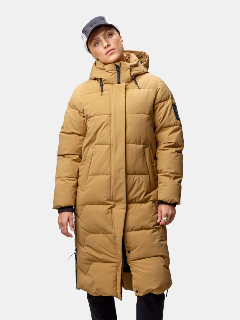 Person, die eine hellbraune Halti Brahe Puffer Long Winterjacke Damen, eine schwarze Hose und eine Baseballmütze vor einem einfarbigen Hintergrund trägt.