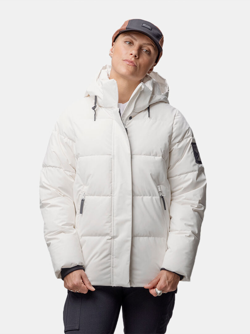 Frau trägt Halti Brahe Puffer Winterjacke aus recyceltem Polyester, schwarze Hose und Two-Tone-Cap vor weißem Hintergrund.