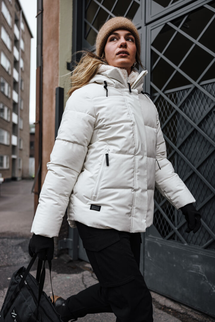 Eine Frau in einer Halti Brahe Puffer Winterjacke Damen geht nach draußen und hält eine schwarze Tasche in einer Hand.