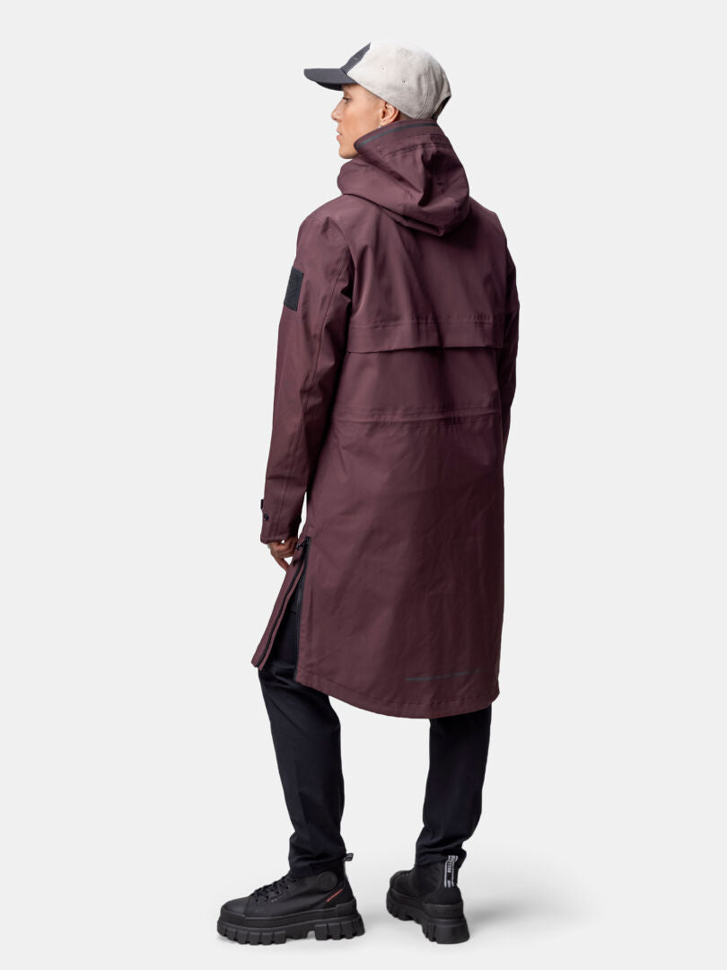 Eine Person in einem langen kastanienbraunen Halti Tokoi II DX Parkajacke Damen, einer schwarzen Hose und einer hellgrauen Mütze, die den Blick abwendet.