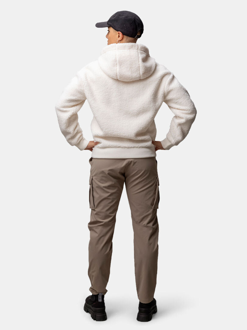 Eine Person in einer Halti Lykky Fleece Jacke Damen, einer hellbraunen Cargohose, schwarzen Schuhen und einer Mütze, die den Blick abwendet.