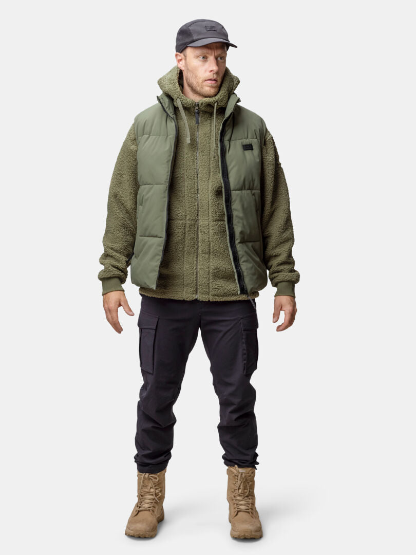 Ein Mann steht vor uns und trägt eine Halti Penger Classic Puffer Weste Herren, eine grüne Fleecejacke, eine schwarze Hose, hellbraune Stiefel und eine schwarze Mütze.
