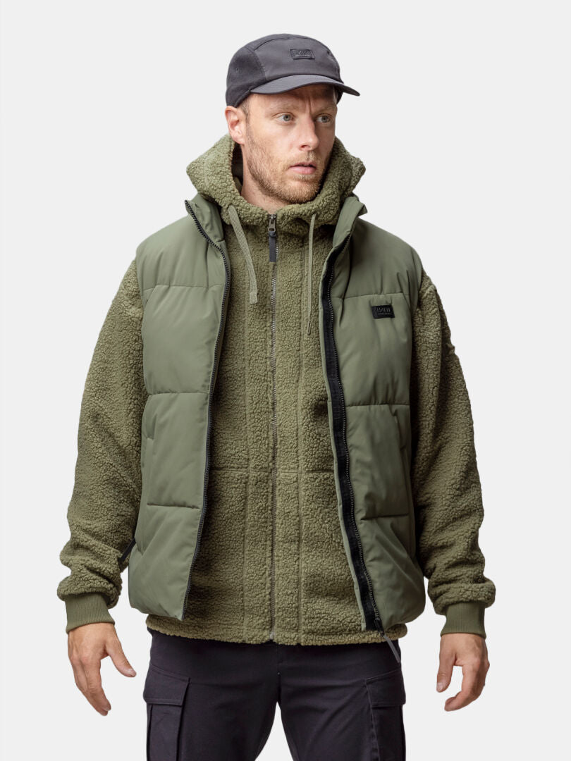 Ein Mann trägt eine grüne Halti Penger Classic Puffer Weste Herren, eine schwarze Cargohose und eine Mütze vor einem weißen Hintergrund.