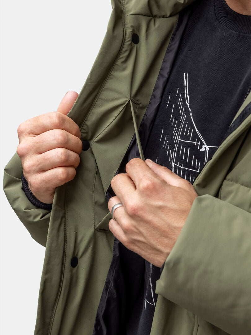 Ein Mann passt den Kordelzug einer Halti Brahe Puffer Winterjacke Herren in Olivgrün über einem schwarzen Grafikshirt an.