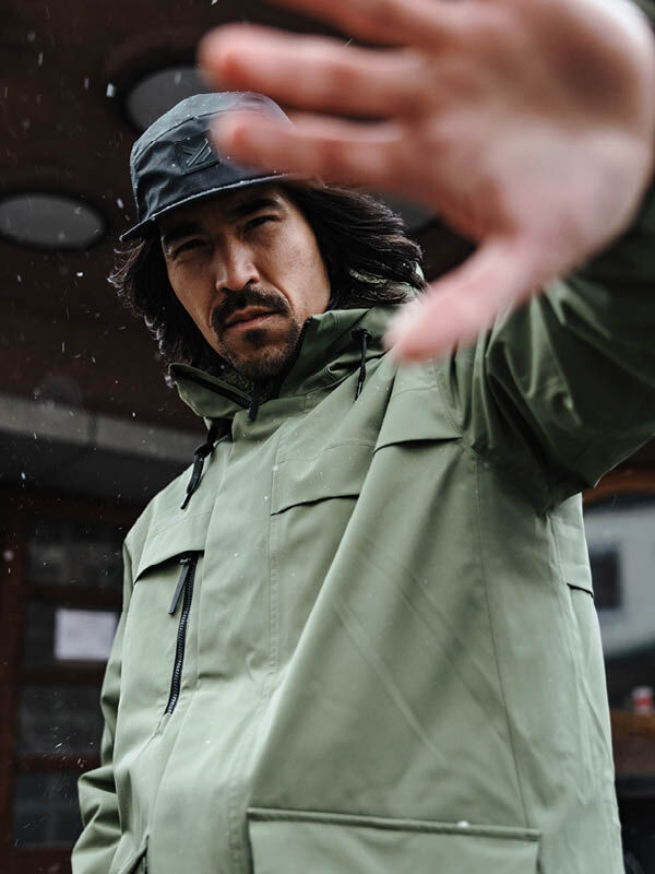 Ein Mann in einer Halti Tokoi II DX Parka Jacke Herren und einem schwarzen Hut hält die Hand hoch und verdeckt damit teilweise sein Gesicht im Freien.