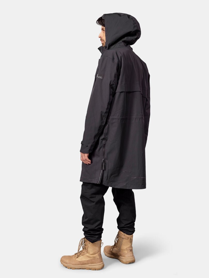 Eine Person, die eine „Halti Tokoi II DX Parka Jacke Herren“ und hellbraune Stiefel trägt, steht seitlich vor einem einfarbigen Hintergrund.