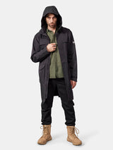Ein Mann in einem „Halti Tokoi II DX“-Parka, einem grünen Shirt, einer schwarzen Hose und hellbraunen Stiefeln vor einem urbanen, hellen Hintergrund.