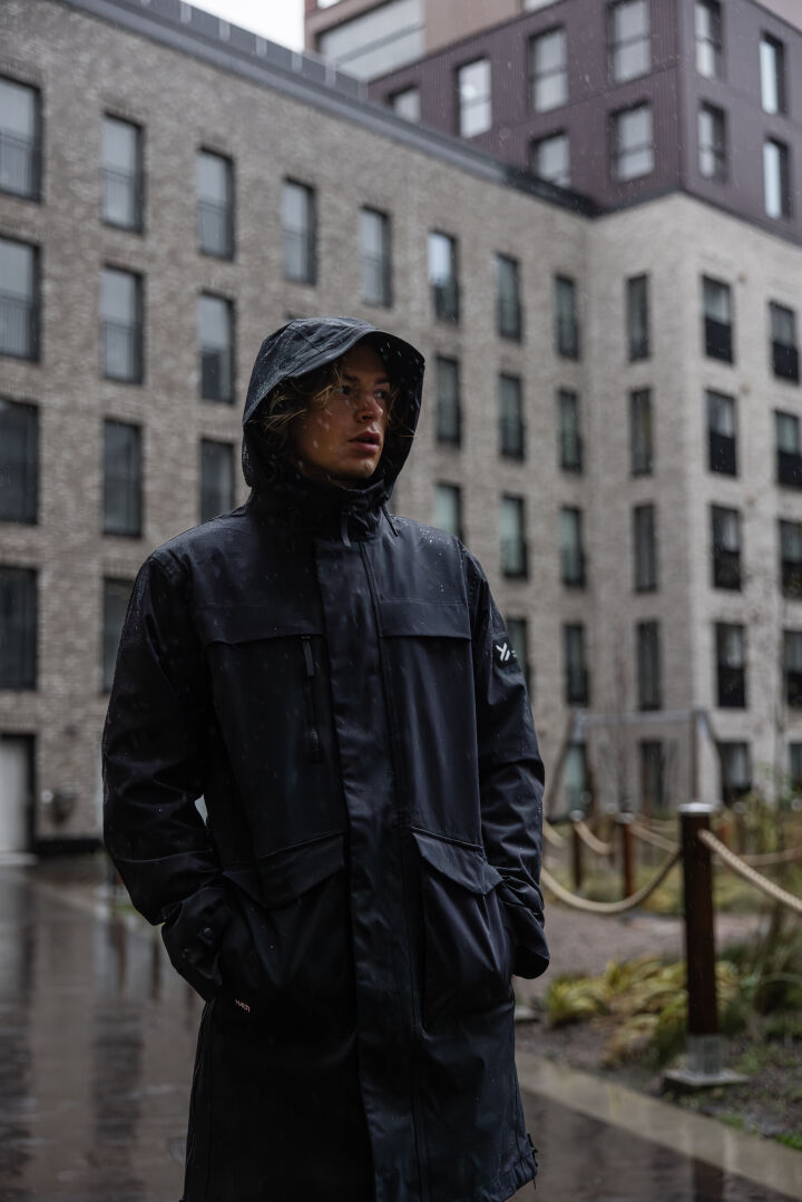 Ein Mann in einem Halti Tokoi II DX Parka steht an einem regnerischen Tag vor einem modernen Gebäude.