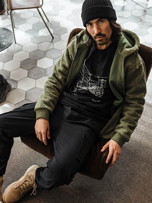 Ein Mann in einer Halti Lykky Fleece Jacke Herren, einer schwarzen Hose und hellbraunen Stiefeln sitzt auf einem Stuhl und blickt in die Kamera.