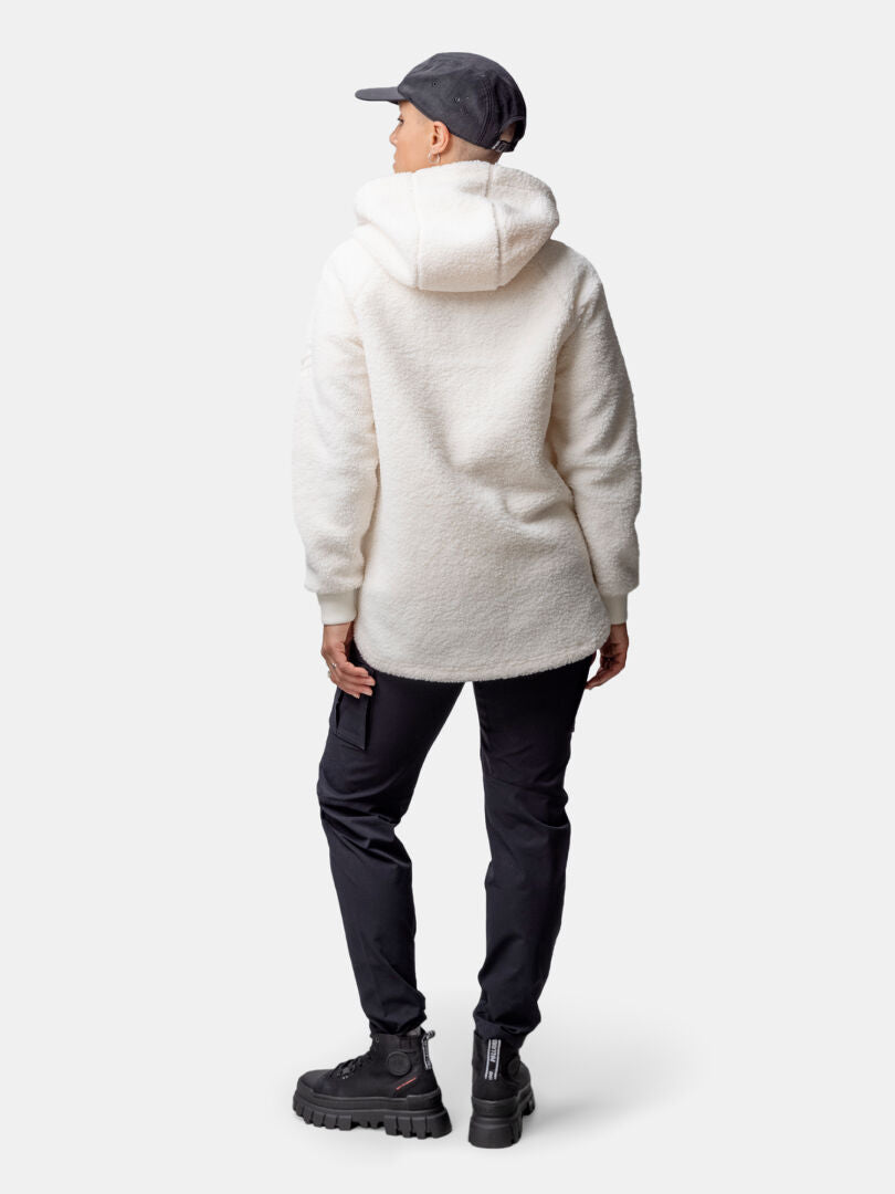 Abgewandte Person in weißem Halti Lykky Fleecepullover Damen, schwarzer Hose, Stiefeln und Mütze vor einfarbigem Hintergrund.