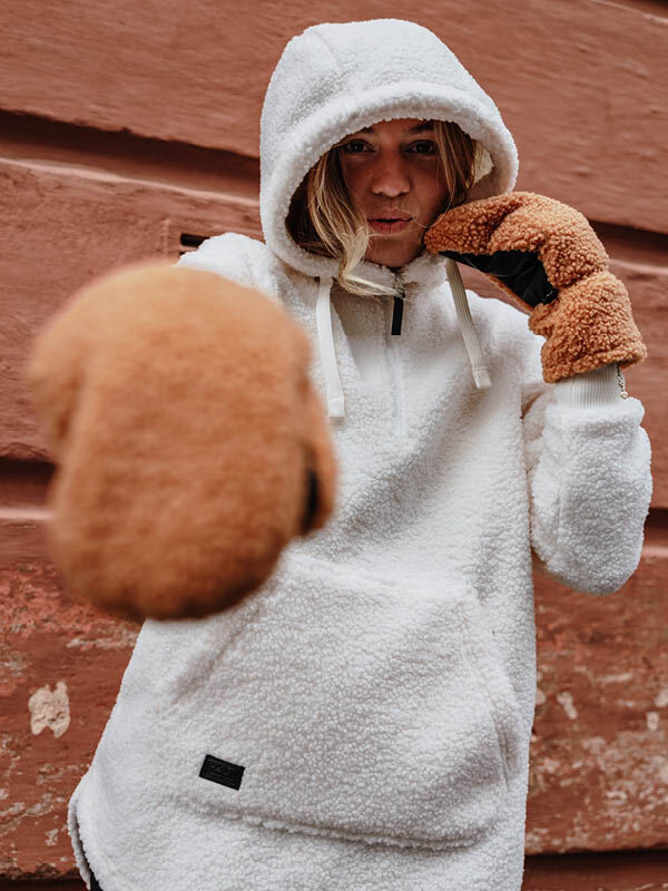 Person in einem Halti Lykky Fleecepullover Damen und braunen Fäustlingen posiert mit erhobenen Fäusten vor einer rot strukturierten Wand.