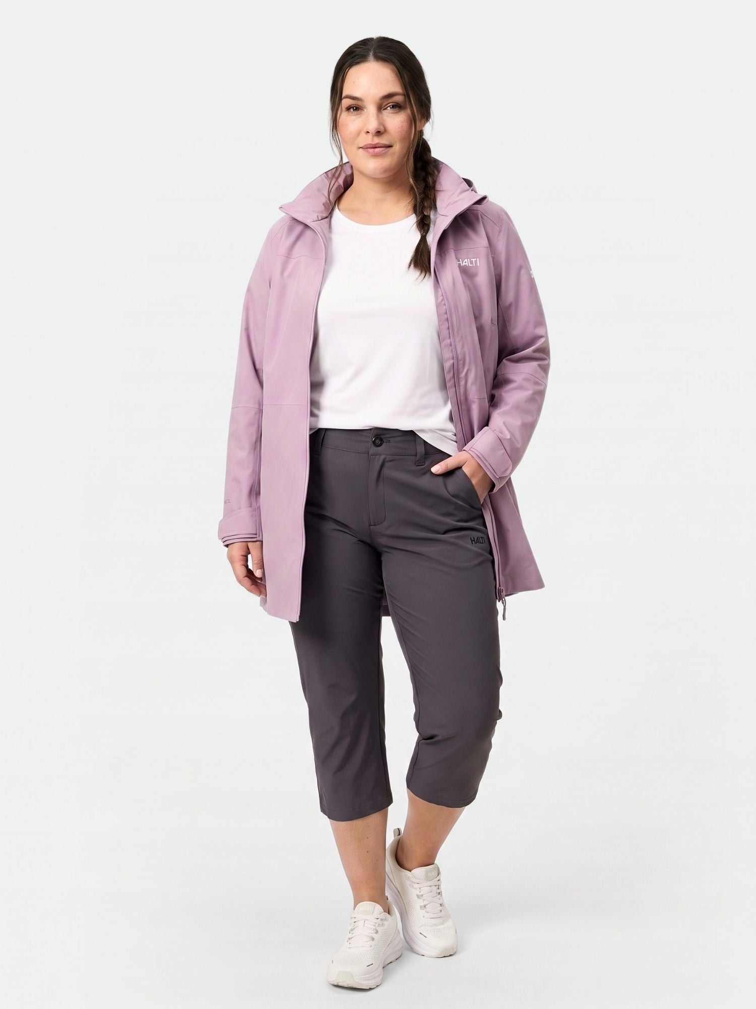 Eine Frau trägt Halti Drive X-stretch Caprihose Damen Plus, eine hellviolette Jacke, ein weißes Hemd und Turnschuhe.
