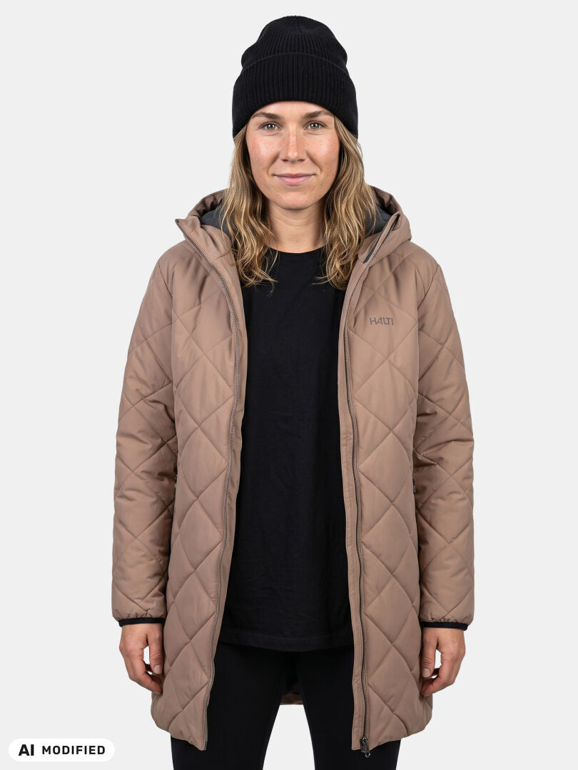 Eine Person in einem hellbraunen „Halti Haapa Steppmantel Damen“, einem schwarzen Shirt und einer schwarzen Mütze vor einem hellen Hintergrund.