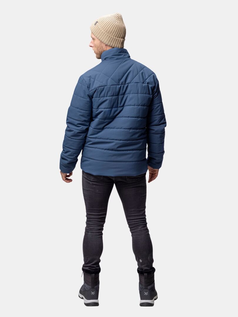 Eine Person in einer blauen Halti Haapa Steppjacke Herren, einer schwarzen Hose, Stiefeln und einer beigen Strickmütze steht mit dem Gesicht zur Seite.