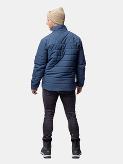 Eine Person in einer blauen Halti Haapa Steppjacke Herren, einer schwarzen Hose, Stiefeln und einer beigen Strickmütze steht mit dem Gesicht zur Seite.