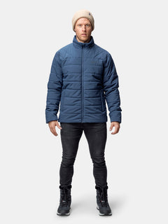 Eine Person in einer blauen Halti Haapa Steppjacke Herren, einer schwarzen Hose, schwarzen Stiefeln und einer beigen Strickmütze sieht nach vorne.