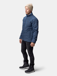 Mann in blauer Halti Haapa Steppjacke Herren, beiger Strickmütze, schwarzer Jeans und Stiefeln auf einfarbigem Hintergrund.