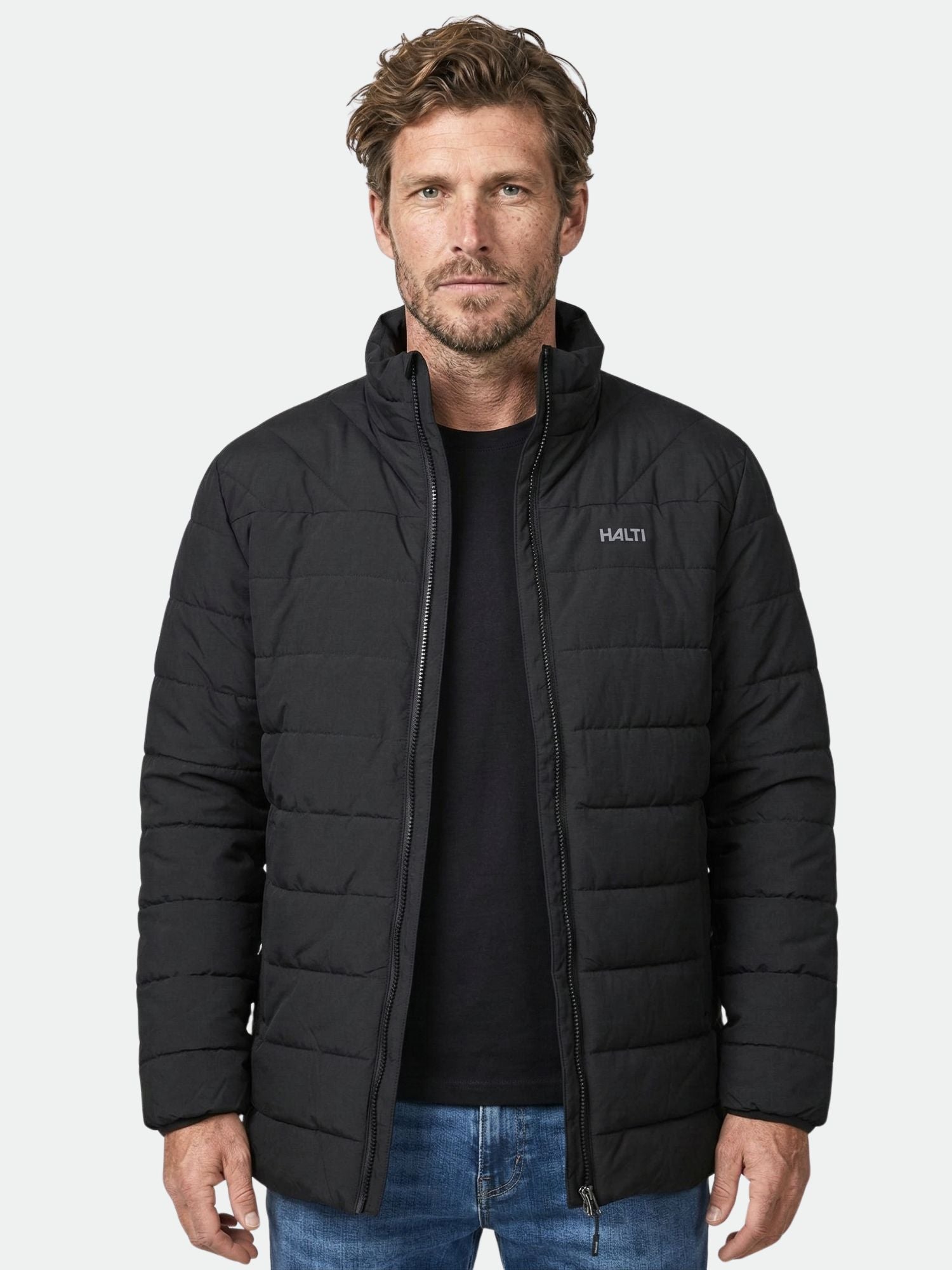 Ein Mann trägt eine schwarze Halti Haapa Steppjacke Herren und blaue Jeans und blickt in die Kamera vor einem schlichten Hintergrund.