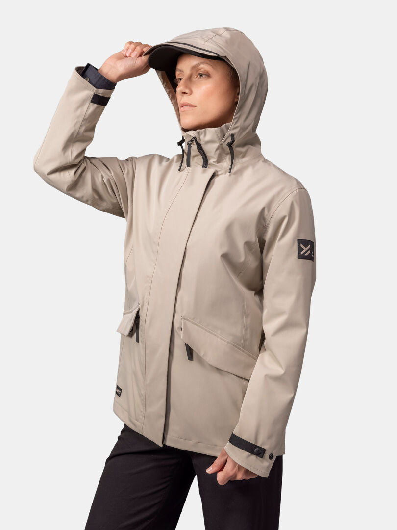 Eine Frau in einer beigen Halti Tokoi DX Jacke Damen und einer schwarzen Hose blickt nach links und hält mit einer Hand die Kapuzenkrempe.