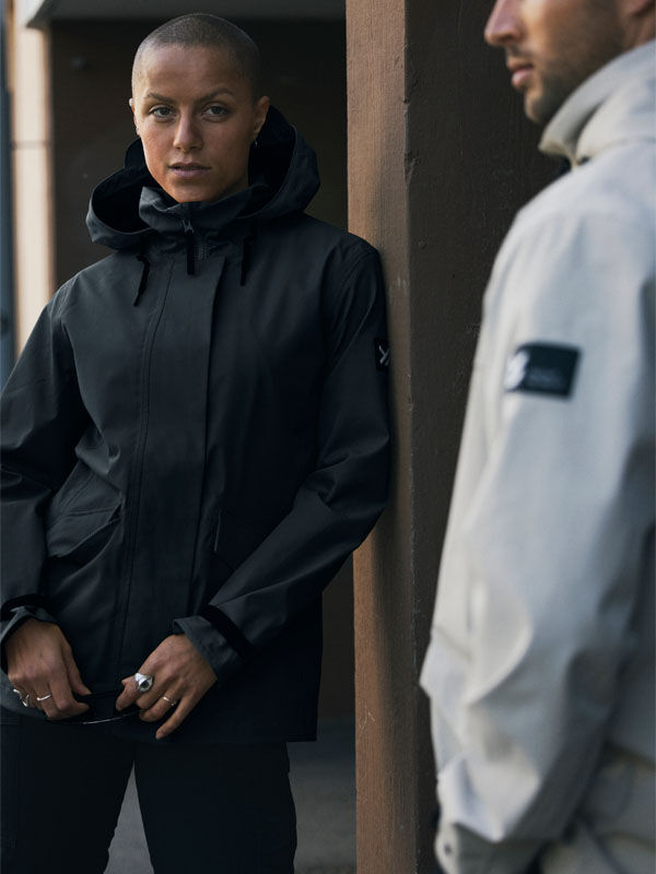 Zwei Personen tragen Halti Tokoi DX Jacke Damen; eine in Schwarz steht vorne, die andere in Hellgrau steht seitlich an einer Wand.