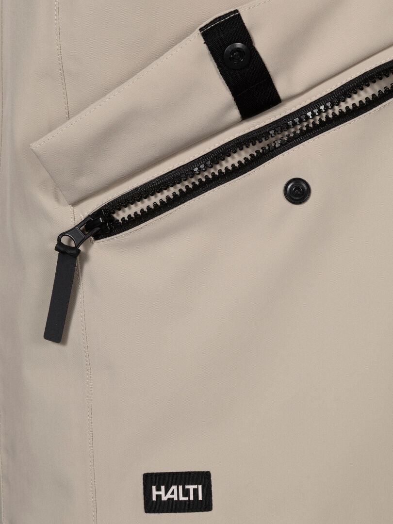 Nahaufnahme der Halti Tokoi DX Jacke Herren in Beige mit Reißverschlusstasche, Druckknopf und schwarzem Halti Logoaufnäher.
