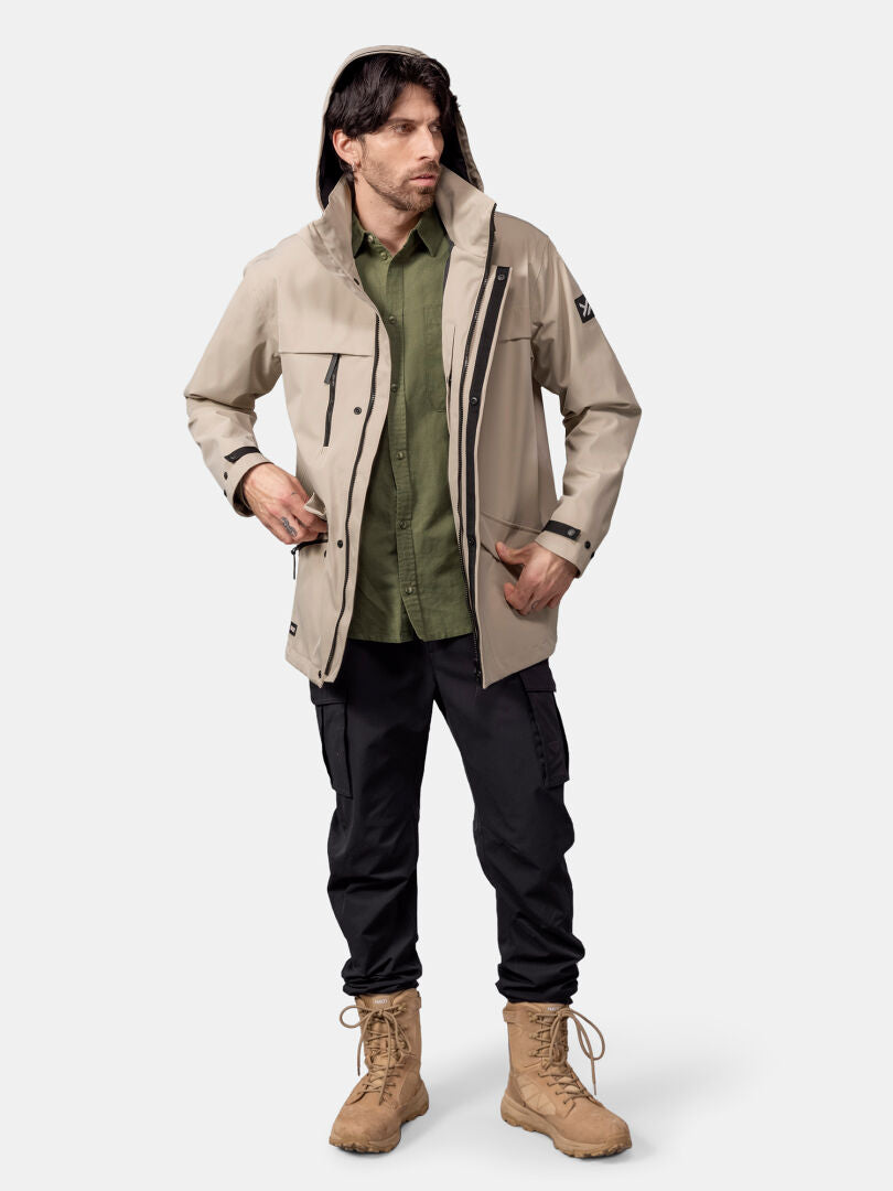 Ein Mann trägt eine beige Halti Tokoi DX Jacke Herren, ein grünes Hemd, eine schwarze Cargohose und hellbraune Stiefel vor einem schlichten Hintergrund.
