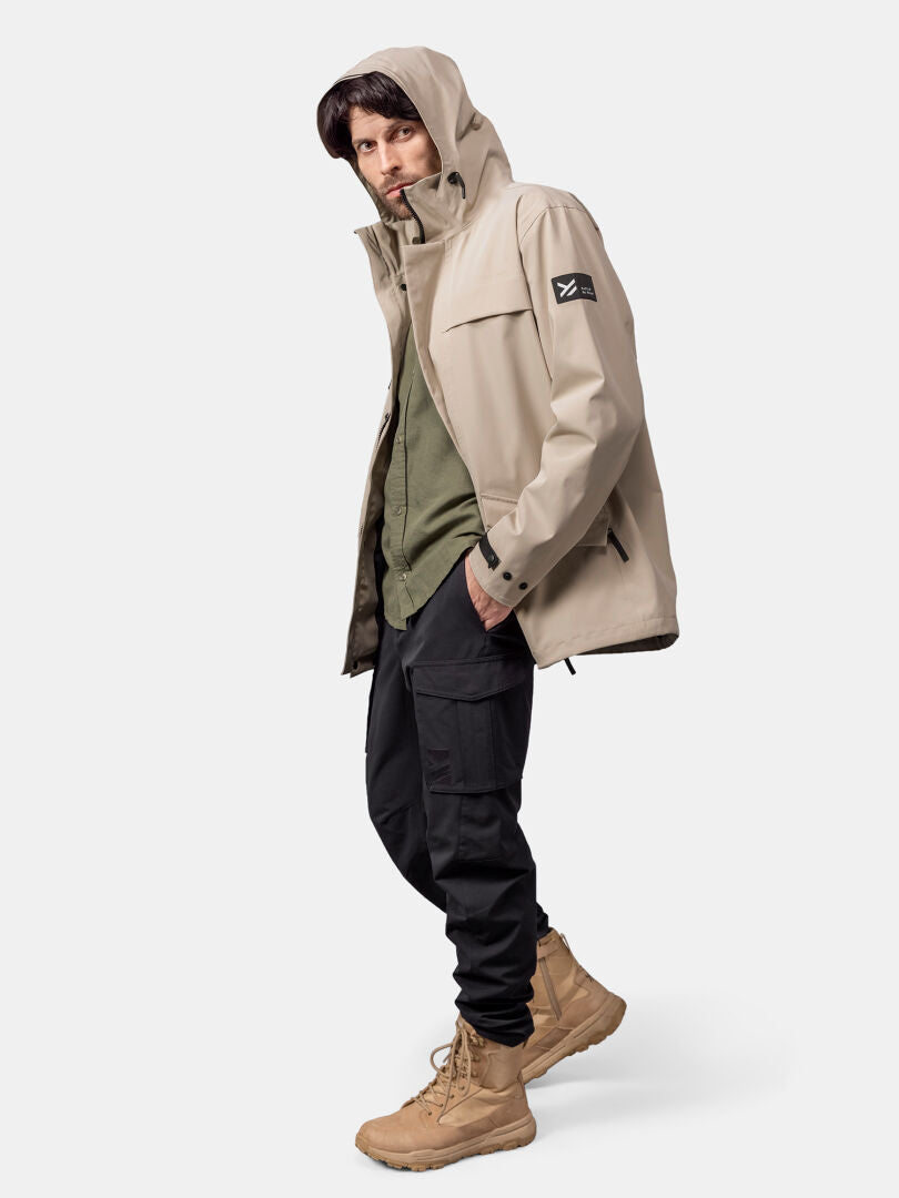 Ein Mann in einer Halti Tokoi DX Jacke Herren, grünem Hemd, schwarzer Cargohose und hellbraunen Stiefeln steht vor einem schlichten Hintergrund.