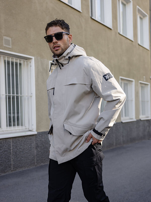 Ein Mann trägt die Halti Tokoi DX Jacke Herren und steht auf dem Gehweg neben einem Gebäude mit vergitterten Fenstern.