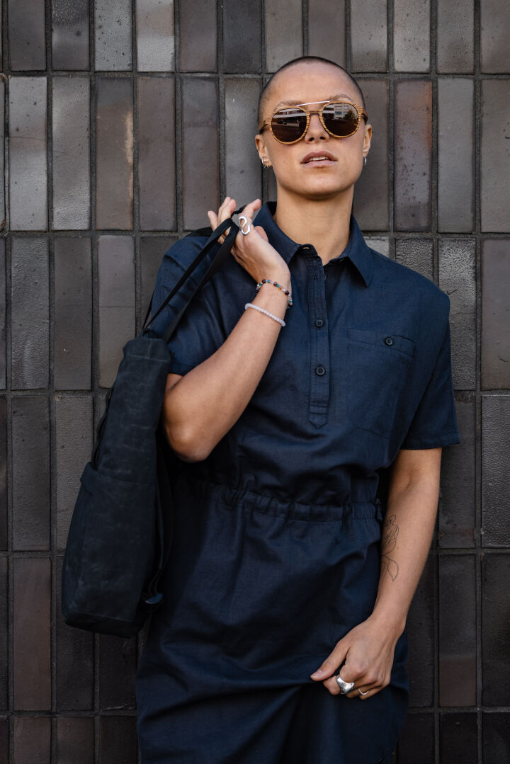 Eine Person in einem dunklen Halti Notsgi Kleid Damen und Sonnenbrille steht an einer gekachelten Wand und hält eine schwarze Tasche.