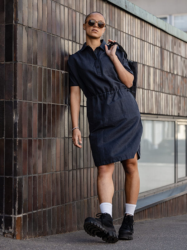 Frau im Halti Notsgi Kleid Damen mit Boots, Sonnenbrille und Tasche lehnt draußen an einer gefliesten Wand.