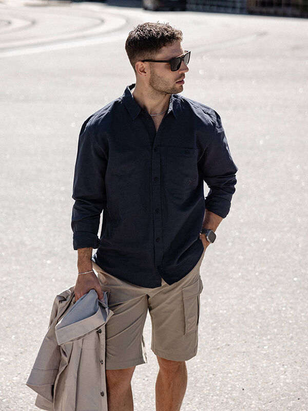 Mann trägt Halti Notsgi Langarm Hemd, Khaki-Shorts, Sonnenbrille, hält helle Jacke im Freien.