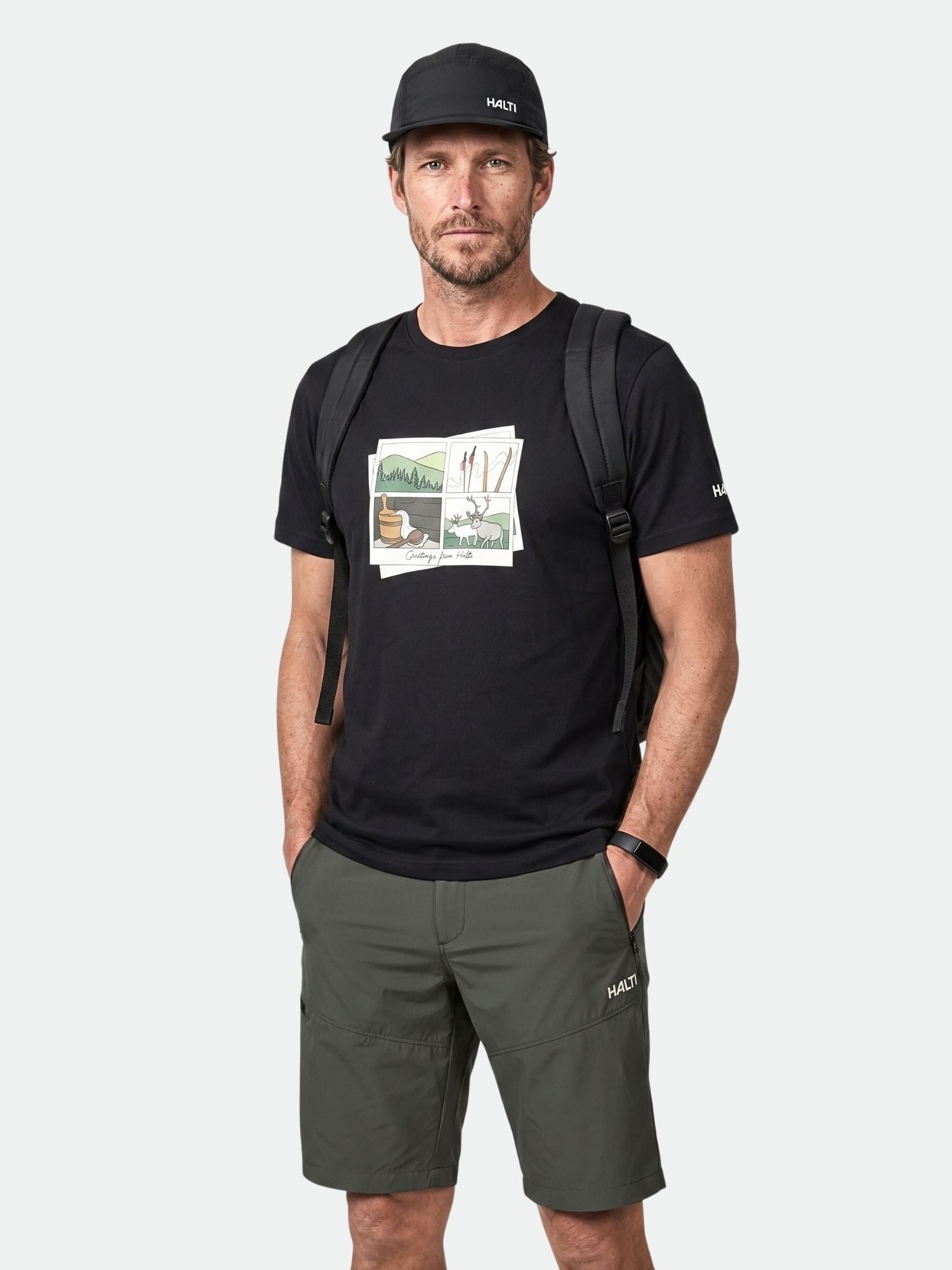 Mann trägt ein schwarzes Halti Matka II T-Shirt Herren, grüne Shorts, Mütze und Rucksack, Hände in den Taschen.