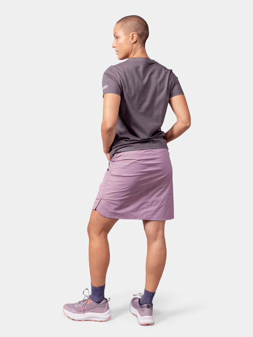 Stehende, abgewandte Person, bekleidet mit Halti Matka II T-Shirt Damen, lilafarbenem Rock, Turnschuhen und marineblauen Socken.