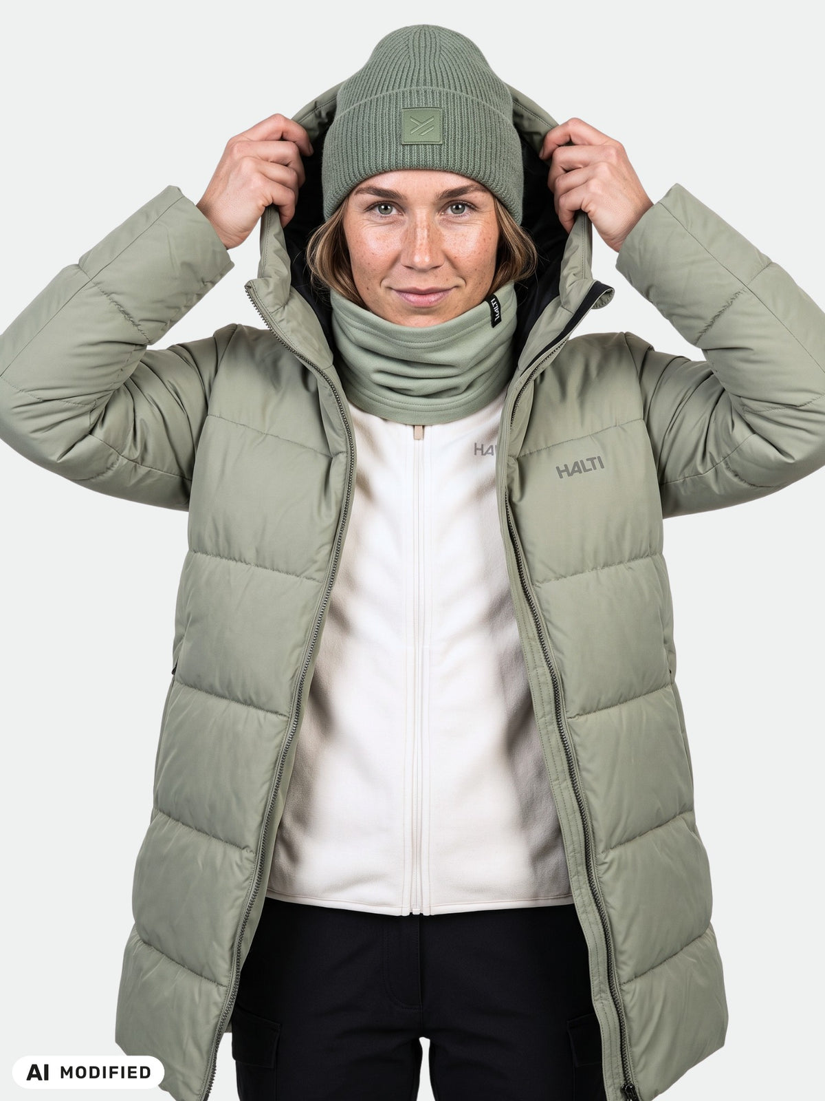 Frau in einem „Halti Leinikko“ Puffer-Parka für Damen, hellgrün, mit hochgezogener Kapuze, vor einem einfarbigen Hintergrund stehend.