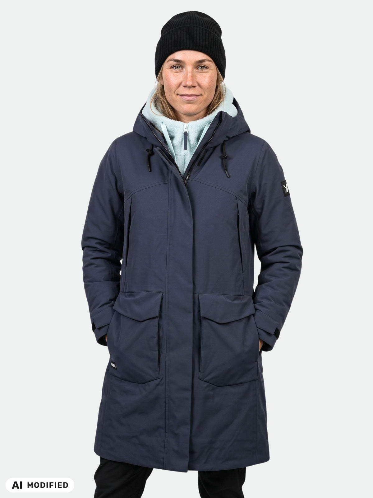 Eine Frau in einem „Halti Kallio DX“-Parka, einem hellen Kapuzenpullover, einer schwarzen Mütze und einer Hose steht vor einem einfarbigen Hintergrund.