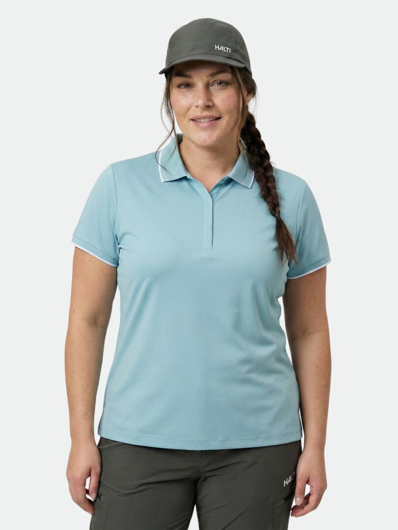 Eine Frau in einem hellblauen Halti Swingi Technisches Poloshirt Damen Plus, einer dunklen Hose und einer grauen Mütze lächelt in die Kamera.