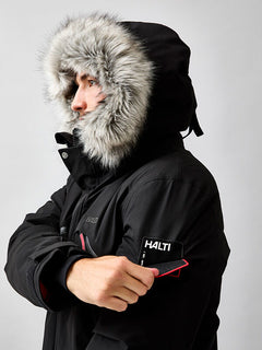 Eine Person in einem schwarzen Halti 1976 Down Parka Unisex mit pelzgefütterter Kapuze trägt einen HALTI-Aufnäher auf dem Ärmel.