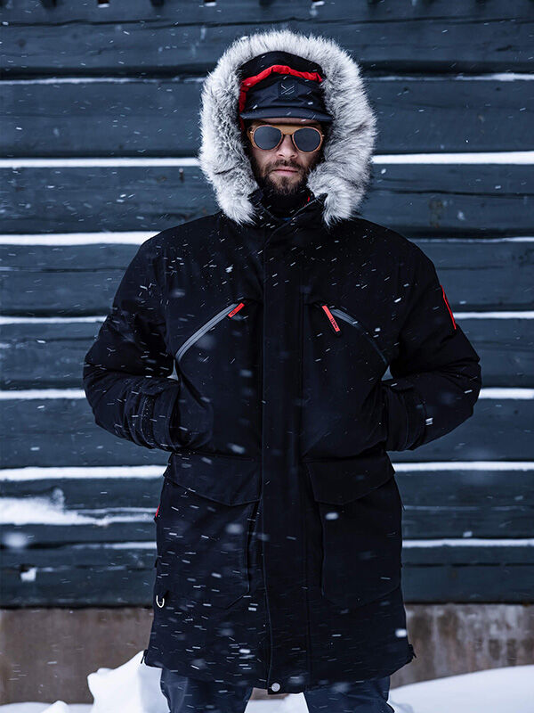 Eine Person in einem Halti 1976 Down Parka Unisex von Halti steht draußen im fallenden Schnee und trägt eine pelzgefütterte Kapuze und Handschuhe.