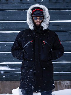 Eine Person, die den Halti 1976 Down Parka Unisex mit pelzgefütterter Kapuze trägt, steht im fallenden Schnee an einer Holzwand.