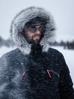 Eine Person im Halti 1976 Down Parka Unisex von Halti steht im Schneegestöber und präsentiert finnisches Design und Sonnenbrillen.