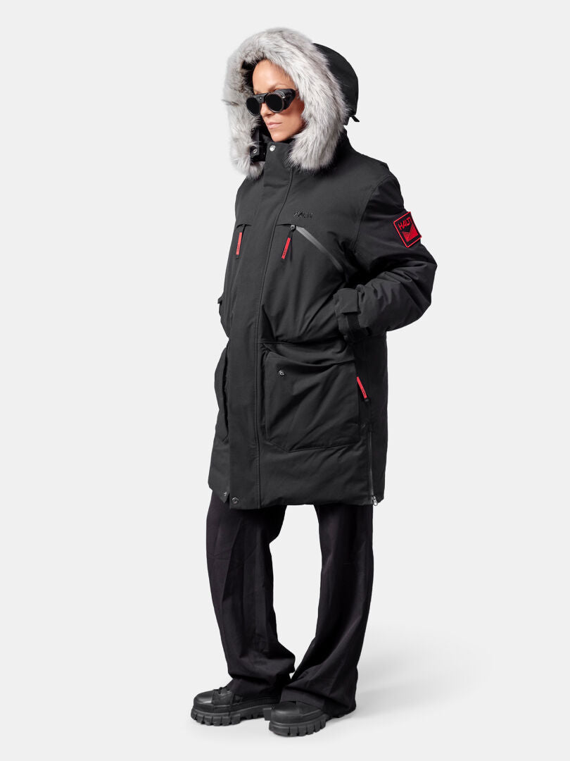 Eine Person trägt einen Halti 1976 Down Parka Unisex von Halti, eine schwarze Hose, Stiefel und eine Sonnenbrille vor einem schlichten Hintergrund.