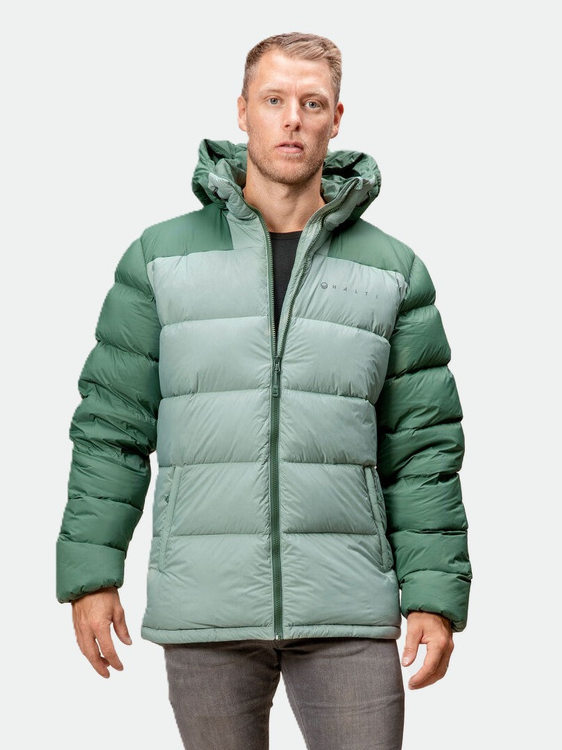 Ein Mann in einer grünen Halti Versant Daunenjacke mit Kapuze, schwarzem Hemd und grauer Hose steht vor einem hellen Hintergrund.