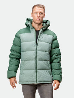 Ein Mann in einer grünen Halti Versant Daunenjacke mit Kapuze, schwarzem Hemd und grauer Hose steht vor einem hellen Hintergrund.