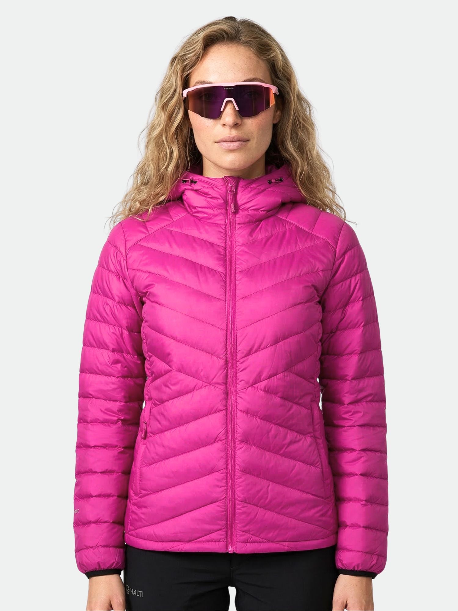 Eine Person trägt eine Halti Huippu Daunenjacke Damen in Rosa und eine große Sonnenbrille vor einem schlichten hellen Hintergrund.