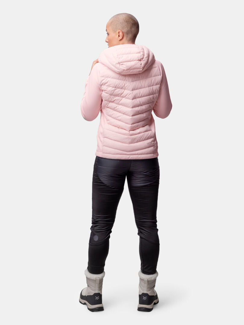 Eine Frau in einer rosa Halti Huippu Hybrid Daunenjacke, einer schwarzen Hose und grauen Stiefeln steht vor einem schlichten Hintergrund.