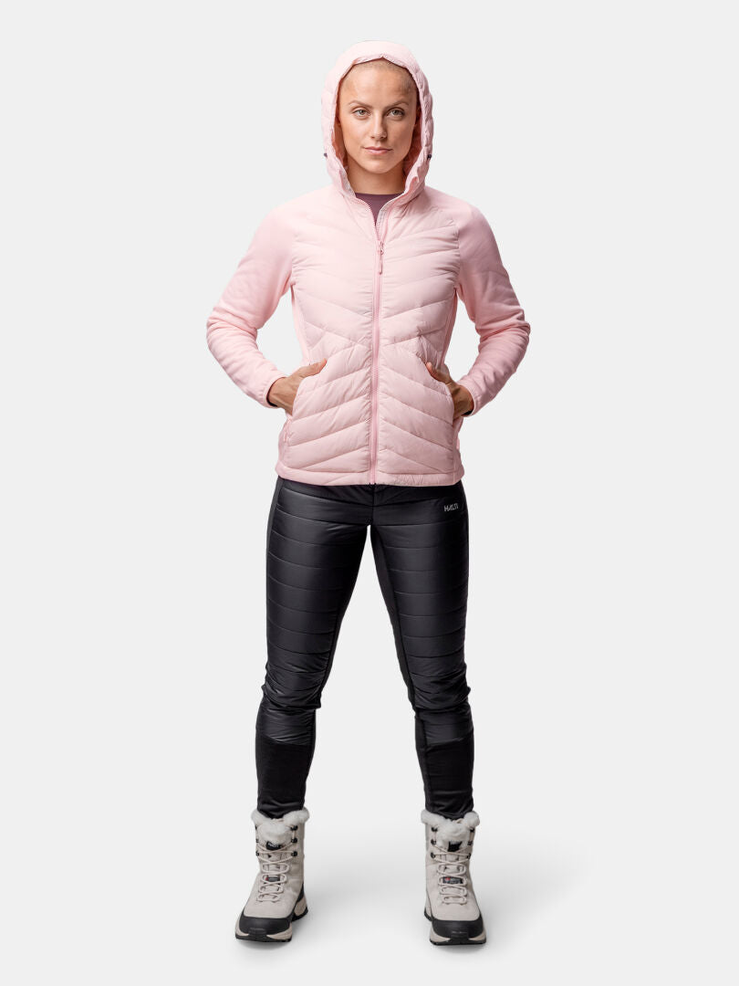 Eine Person trägt eine rosafarbene Halti Huippu Hybrid Daunenjacke Damen, eine schwarze Hose und graue Winterstiefel vor einem schlichten Hintergrund.