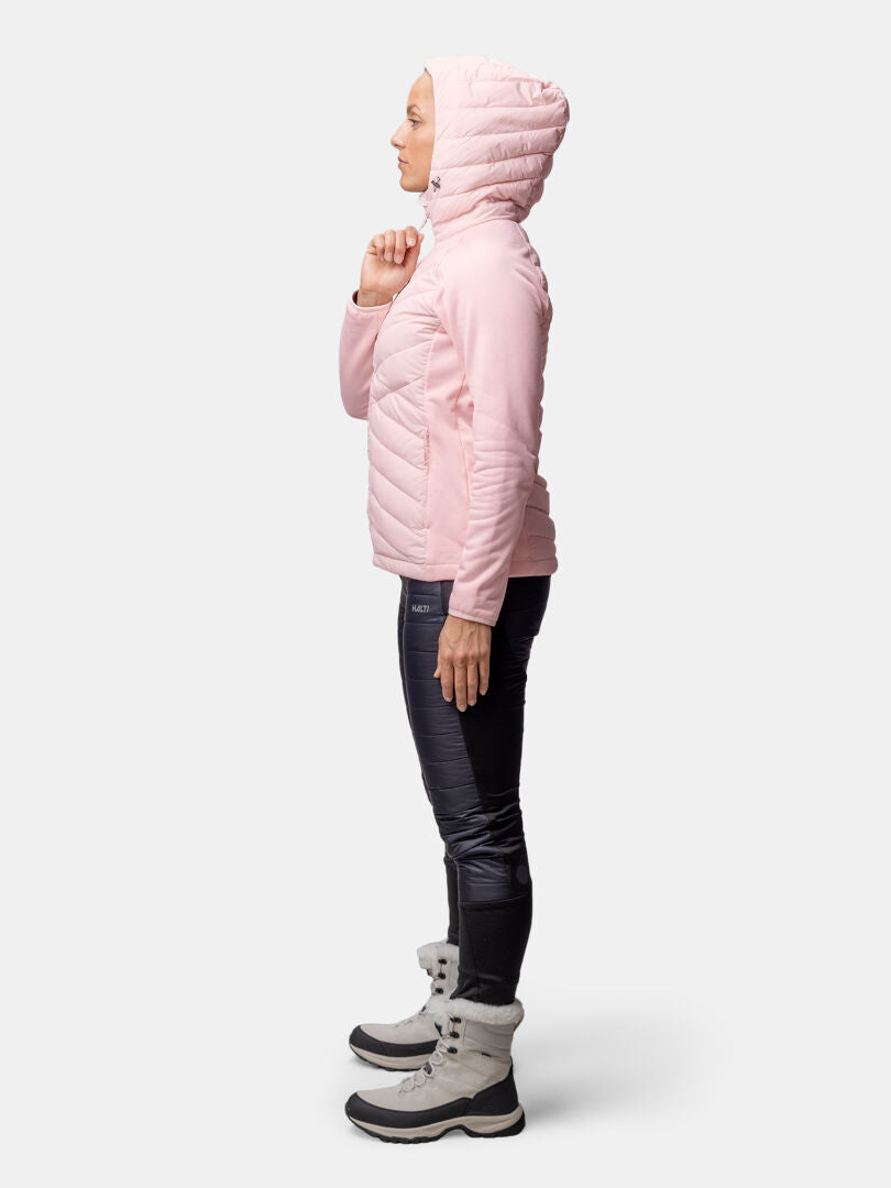 Eine Person in einer rosa Halti Huippu Hybrid Daunenjacke Damen mit Pertex Quantum Ripstop steht im Profil vor einem einfarbigen Hintergrund.