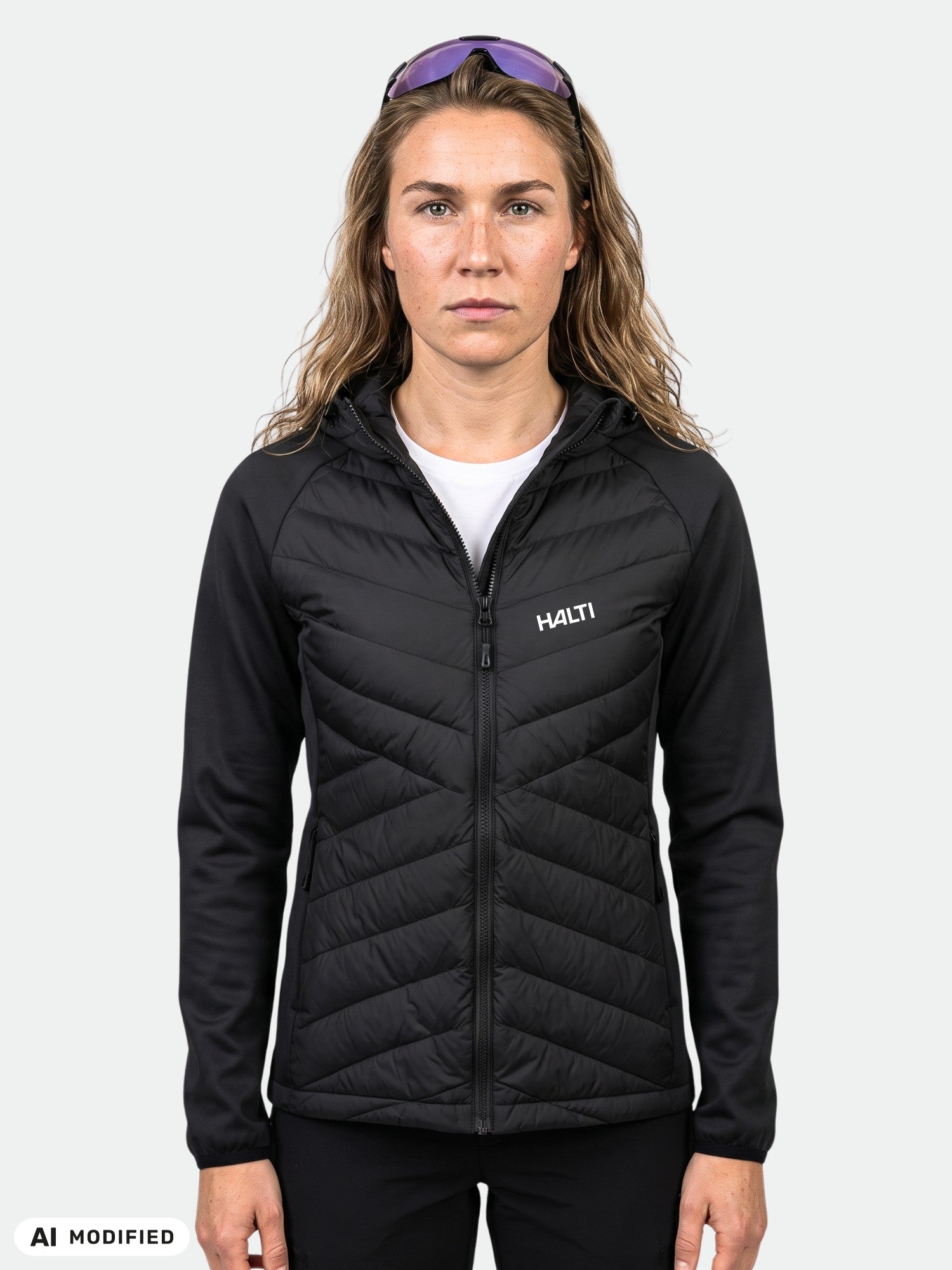 Eine Person, die eine schwarze Halti Streams Hybrid Midlayer Jacke für Damen und eine Sonnenbrille trägt und vor einem einfarbigen hellen Hintergrund steht.